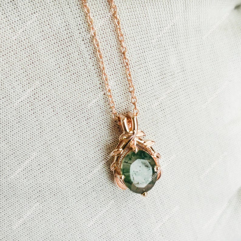 May include: Rose gold necklace featuring a round, green gemstone pendant. The pendant is set in a decorative frame with leaf-like accents. The chain is delicate and complements the pendant's design. The jewelry is displayed on a textured, off-white surface.