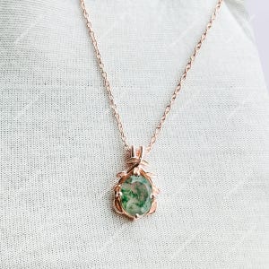 May include: A rose gold necklace featuring a pendant with a green and white gemstone. The pendant is teardrop-shaped and surrounded by a decorative rose gold frame. The necklace chain is delicate and hangs on a white background.