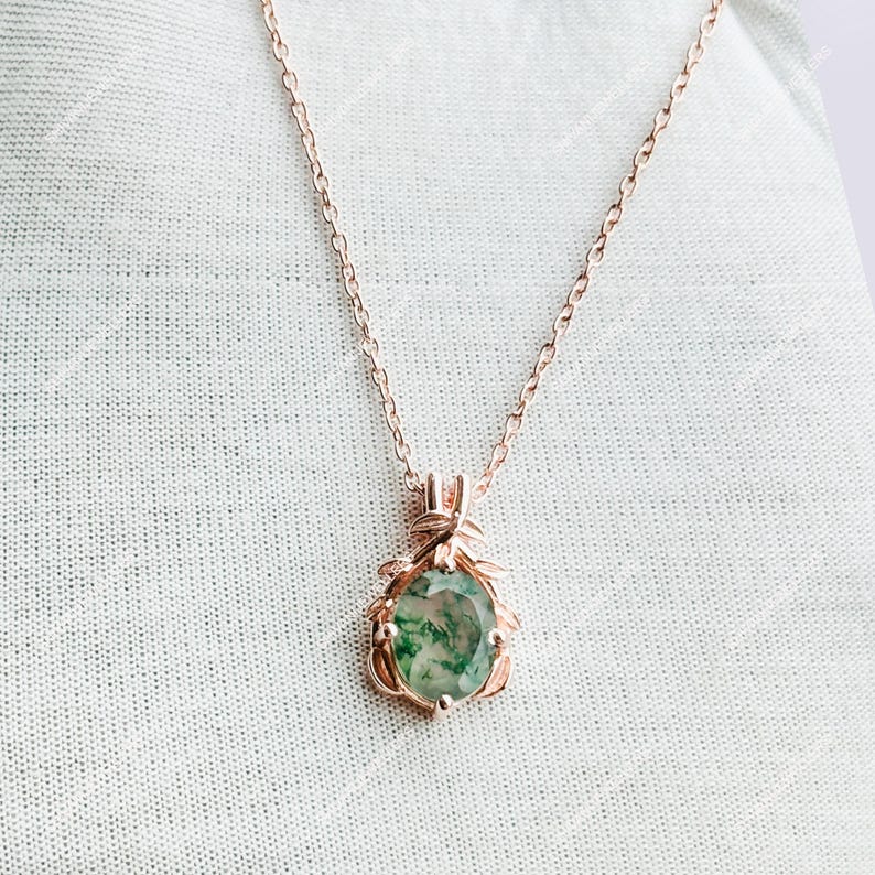 May include: A rose gold necklace featuring a pendant with a green and white gemstone. The pendant is teardrop-shaped and surrounded by a decorative rose gold frame. The necklace chain is delicate and hangs on a white background.