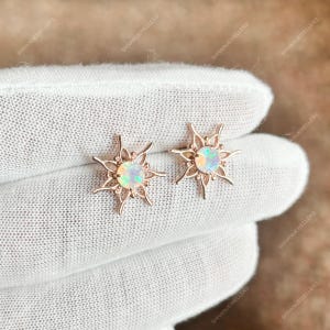 Könnte beinhalten: Ein Paar roségoldene Ohrstecker. Jeder Ohrring hat einen zentralen Opalstein, der von einem Sonnenstrahl-Design umgeben ist. Die Opale zeigen ein Farbspiel mit grünen, blauen und gelben Blitzen. Die Ohrringe werden auf einer weißen Oberfläche präsentiert.