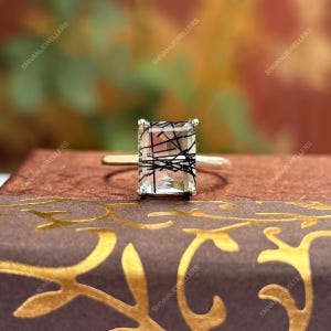 Pode incluir: Um anel de ouro com uma pedra preciosa retangular e transparente com inclusões pretas. O anel está sobre uma superfície castanha com elementos decorativos dourados.