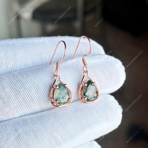 Op de afbeelding: Een paar roségouden oorhangers. Elke oorbel heeft een druppelvormige groene edelsteen in een delicaat montuur. De oorbellen worden gepresenteerd op een wit oppervlak.
