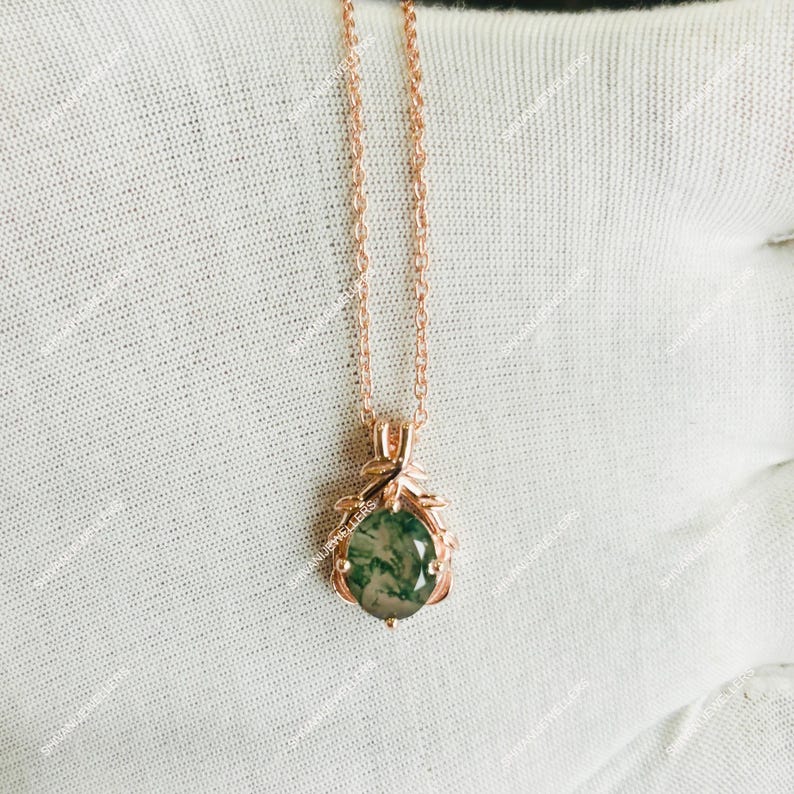 May include: A rose gold necklace featuring a pendant with a green moss agate stone. The pendant is teardrop-shaped and surrounded by a decorative rose gold setting. The chain is delicate and hangs on a white surface.