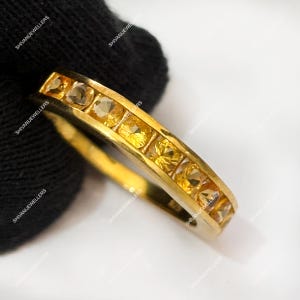Puede incluir: Un anillo de oro con una fila de piedras preciosas amarillas de talla cuadrada. El anillo tiene un acabado pulido y está sobre un fondo negro. Las piedras preciosas son una mezcla de amarillo claro y oscuro.