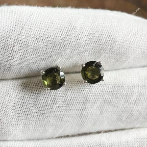 Puede incluir: Un par de pendientes de botón redondos de piedra preciosa verde oliva con postes de plata. Los pendientes están colocados sobre un fondo blanco texturizado. Las piedras preciosas tienen un aspecto facetado.