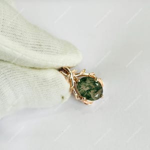 May include: A rose gold pendant featuring an oval-cut moss agate gemstone. The pendant has a leaf-like design around the stone and a bail for a chain. The gemstone has green and white inclusions.