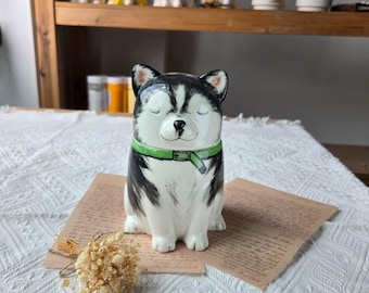 May include: A ceramic husky dog figurine with black and white fur, a green collar, and closed eyes. The figurine is sitting on a piece of paper with text. Other ceramic animal figurines are visible in the background.
