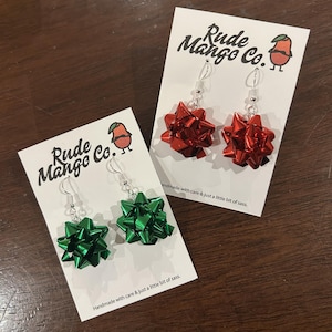 Puede incluir: Dos pares de pendientes en forma de lazo, uno rojo y otro verde, expuestos en tarjetas blancas. Las tarjetas muestran el texto "Rude Mango Co." y "Handmade with care & just a little bit of sass."