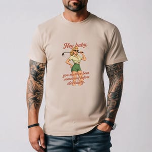 May include: Beige t-shirt featuring a retro illustration of a woman in golf attire holding a golf club. The text on the shirt reads "Hey baby, you must've been something before electricity."