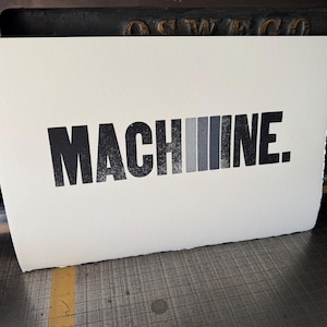May include: A rectangular white paper print featuring the black text "MACH" followed by a series of four vertical bars in a gradient from light grey to black, and the black text "INE." with a full stop at the end, all in a bold sans-serif font.