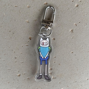 May include: A clear acrylic keychain featuring a cartoon character with a white hat, blue shirt, and blue shorts. The keychain has a silver-colored metal clasp for attaching to keys or bags. The character has a smiling expression.