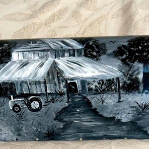 Puede incluir: Un buzón pintado a mano con un paisaje rural en tonos grises y blancos. La obra de arte representa una granja, un tractor, árboles y un camino sinuoso. El buzón es de metal.