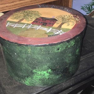 Puede incluir: Una caja decorativa redonda pintada a mano, con un exterior verde y una tapa que presenta una escena rural. La tapa muestra un granero rojo, una valla blanca y &aacute;rboles, con la palabra "Welcome" pintada en la parte superior. La caja tiene un borde rojo oscuro.