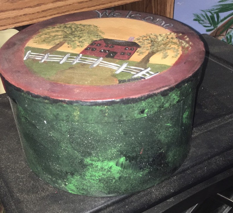 Puede incluir: Una caja decorativa redonda pintada a mano, con un exterior verde y una tapa que presenta una escena rural. La tapa muestra un granero rojo, una valla blanca y &aacute;rboles, con la palabra "Welcome" pintada en la parte superior. La caja tiene un borde rojo oscuro.