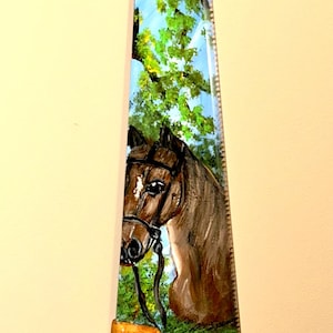 Puede incluir: Hoja de sierra pintada a mano con la cabeza de un caballo marrón con brida, sobre un fondo de follaje verde y cielo azul. El mango de madera está asegurado con remaches de latón, creando una pieza decorativa única.