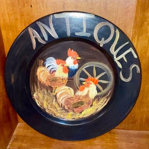 Puede incluir: Un plato redondo negro con la palabra "ANTIQUES" pintada en blanco alrededor del borde. El centro del plato presenta una escena pintada a mano de tres gallos en un entorno de granero.