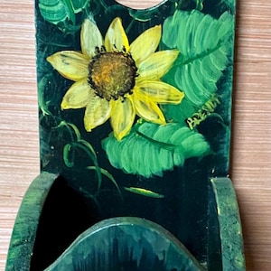 Puede incluir: Un portacartas de pared de madera pintado de negro con un girasol y hojas verdes. El portacartas tiene una pequeña abertura en la parte superior y una ranura para el correo.