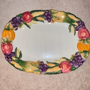 May include: White oval ceramic serving platter with a raised border decorated with a colorful autumn harvest theme. The border features grapes, apples, corn, and pumpkins.