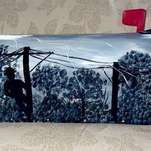 Puede incluir: Un buzón pintado a mano con una escena de paisaje rural. El diseño presenta una figura silueteada, árboles y postes de servicios públicos en tonos grises y azules. El buzón tiene una bandera roja.