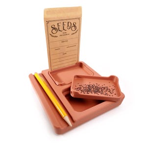 May include: A terracotta-colored seed starting kit with a paper seed packet labeled "SEEDS" and a yellow pencil. The kit includes a tray with multiple compartments and a small tray filled with seeds. The seed packet has space for seed type, variety, and notes.