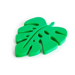 May include: A bright green, leaf-shaped coaster with a monstera design. The coaster has a smooth surface and several cut-out holes. The leaf is a vibrant green color and sits on a white background.