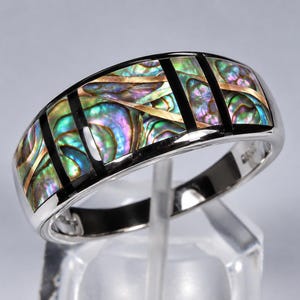 Op de afbeelding: Een zilverkleurige ring met een brede band ingelegd met iriserende abalone schelpsegmenten. De schelp vertoont levendige groene, blauwe en paarse tinten, gescheiden door dunne, verticale zwarte strepen en smalle goudkleurige metalen accenten.