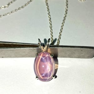Pode incluir: Um colar de prata com um pingente. O pingente apresenta uma grande pedra preciosa rosa oval em forma de estrela, engastada em uma moldura de prata. A pedra preciosa tem um efeito de estrela visível. A corrente é delicada e prateada.