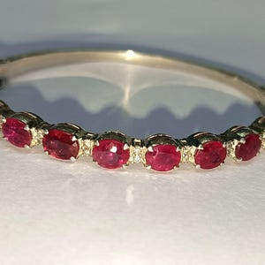 Könnte beinhalten: Ein silbernes Armband mit einer Reihe ovaler, leuchtend roter Edelsteine. Die Edelsteine sind in einzelnen Fassungen gefasst, und das Armband hat ein klassisches, elegantes Design. Der Verschluss ist auf der linken Seite sichtbar.