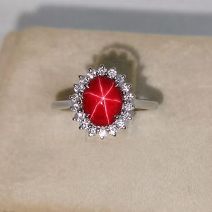 Könnte beinhalten: Ein silberfarbener Ring mit einem zentralen, ovalen roten Edelstein, der ein markantes sechsstrahliges Sternmuster aufweist. Der rote Stein ist von einem Kranz aus kleinen, klaren, rund geschliffenen Steinen in einer gewellten Fassung umgeben.