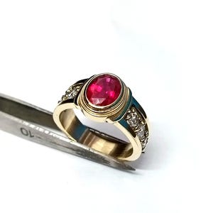 May include: A gold ring with a large oval ruby gemstone as the centerpiece. The ring has a detailed design with smaller accent stones and is held by a silver tool against a white background. The ruby is a vibrant red color.