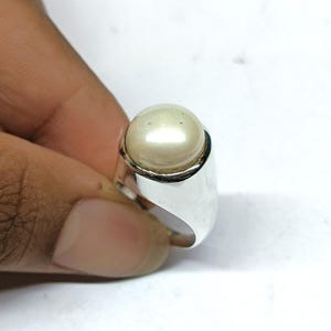 Puede incluir: Un anillo de plata con una perla grande, redonda y de color crema. El anillo tiene una banda lisa y pulida y un engaste de bisel que sujeta la perla de forma segura. Una joya clásica.
