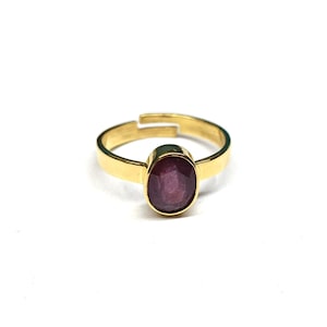 Puede incluir: Un anillo ajustable de color dorado con una gema ovalada de color rojo intenso. La banda del anillo tiene un diseño sencillo y pulido, y la gema está engastada en un bisel. El anillo se muestra sobre un fondo blanco.