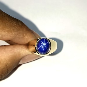 Puede incluir: Anillo de oro con una gran gema ovalada de color azul oscuro con un patrón en forma de estrella. El anillo tiene una banda de oro pulido y un diseño clásico. La gema es de un azul intenso.