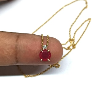 May include: A gold necklace with a round, deep red gemstone pendant. A small, clear stone sits above the red gem. The pendant hangs from a delicate gold chain. The background is white.