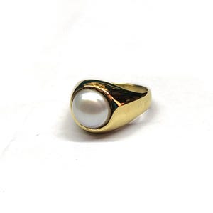 Könnte beinhalten: Ein goldener Ring mit einer großen, runden, weißen Perle. Der Ring hat ein dickes, poliertes Goldband, das sich um die Perle wölbt. Die Perle ist zentriert und scheint sicher im Gold gefasst zu sein. Das Design ist glatt und elegant.