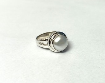 May include: A silver ring featuring a large, round, pearlescent white pearl. The ring has a thick band with a split design and a decorative, layered setting around the pearl. The ring is set against a white background.