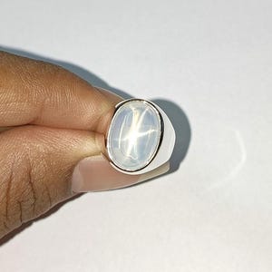 May include: A silver ring with a large, oval, pale gemstone displaying a star effect. The ring has a plain, polished band and a bezel setting. The gemstone is translucent, with a soft glow. The ring is held in a hand.
