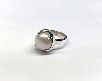 May include: A silver ring with a large, round, lustrous pearl. The pearl is held by four prongs, and the band is polished silver. The ring is set against a white background. A classic piece of jewellery.
