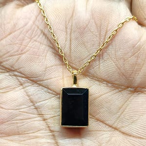 Peut inclure: Un collier doré avec un pendentif rectangulaire noir. Le pendentif est serti dans un cadre doré et suspendu à une fine chaîne dorée. Le collier est présenté sur un fond clair.