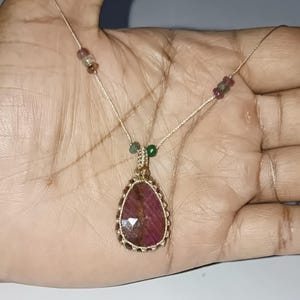 以下が含まれることがあります： 手作りのネックレス。涙の形をした、ファセット加工された、濃い赤色の宝石のペンダントが特徴です。ペンダントは金色の織りフレームにセットされ、繊細なベージュ色のコードに吊り下げられています。小さなカラフルなビーズがコードを飾っています。