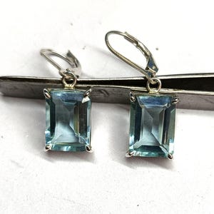 Puede incluir: Un par de pendientes de plata con cierre de palanca, cada uno con una piedra preciosa rectangular de color azul claro. Las piedras están engastadas en un marco de plata, creando un aspecto clásico y elegante. Los pendientes se muestran sobre un fondo neutro.