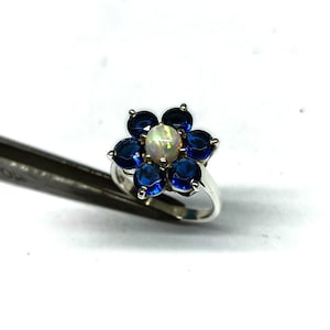 May include: A silver ring featuring a flower design. The center stone is an oval opal, surrounded by six round, deep blue sapphires. The ring is set against a white background, showcasing the vibrant colors of the gemstones.
