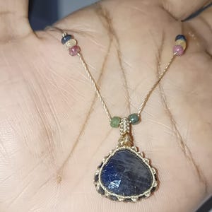 May include: A handcrafted necklace featuring a large, faceted, dark blue gemstone pendant. The pendant is intricately wrapped with a gold-colored wire and suspended from a delicate, beige cord. Small, colorful beads accent the cord near the clasp.