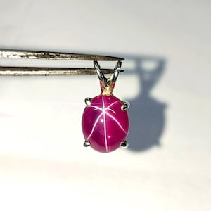 Peut inclure: Un pendentif en rubis ovale vif avec un effet d'étoile. La pierre précieuse rose foncé est sertie dans un métal argenté avec une petite boucle pour une chaîne. Le pendentif est tenu par une pince sur fond blanc.