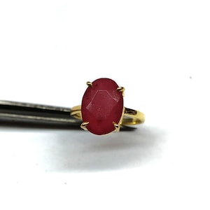May include: A gold ring with a large, oval-cut ruby gemstone. The ruby is a deep red colour and is held in place by four prongs. The ring's band is a simple, polished gold.
