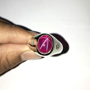 May include: A silver ring with a large, oval, deep pink gemstone featuring a six-ray star pattern. Two small diamonds are set on either side of the main stone. The ring is a classic signet style.
