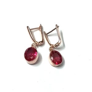 May include: A pair of rose gold earrings with oval, deep red gemstones. The earrings have a lever back closure and a polished finish. The gemstones are set in a bezel setting, and the earrings are displayed on a white background.