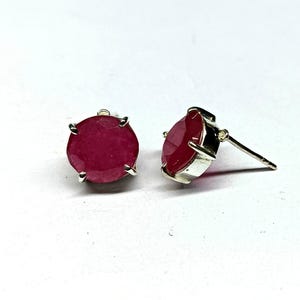 Könnte beinhalten: Ein Paar Ohrstecker mit runden, tiefroten Edelsteinen, die in silberfarbenem Metall gefasst sind. Die Edelsteine haben eine lebendige, gesättigte Farbe, und die Ohrringe haben ein klassisches, minimalistisches Design. Die Ohrringe werden auf weißem Hintergrund gezeigt.