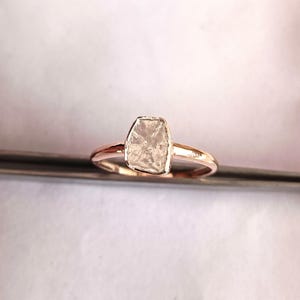 May include: A rose gold ring with a large, rectangular, raw-cut diamond. The diamond has an irregular shape and a clear, crystalline appearance. The band is polished, in a warm rose gold colour.
