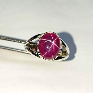 May include: A silver ring featuring an oval, deep red gemstone with a star-like pattern. The ring's band has a split design, and the gem is held in a bezel setting. The ring is displayed against a white background.